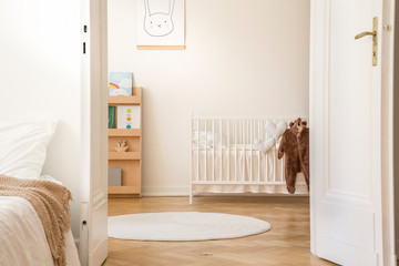 Stylish bedroom interior with open door to scandinavian nursery with white crib, poster on the wall...