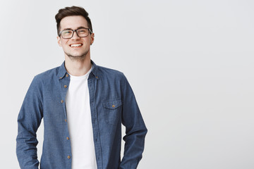 Waist-up shot of happy friendly-looking handsome young smart male coworker in glasses and blue shirt smiling standing over gray background proud and satisfied, grinning at camera from joy