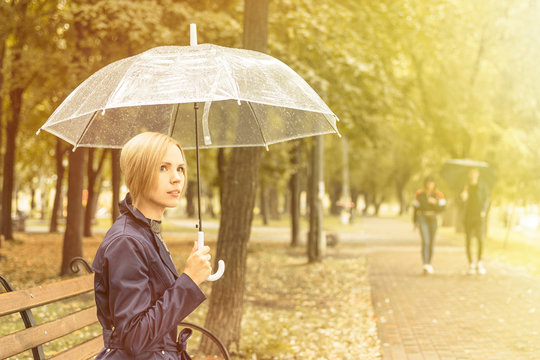 A woman in a raincoat with a transparent umbrella is sitting on a bench. Toned.