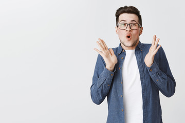 Enthusiastic happy and excited young handsome man with bristle in glasses and blue shirt raising hands in joy, clapping palms folding lips hearing awesome news feeling delighted and surprised