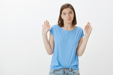 Not my fault, I am innocent. Unaware careless charming european woman in blue t-shirt, raising palm in surrender and smirking at camera, wanting to reject offer and giving up over grey wall