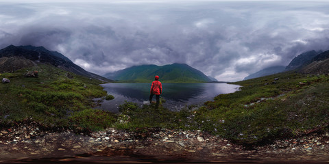 man stands by lake in red jacket in cloudy weather. Spherical panorama 360vr