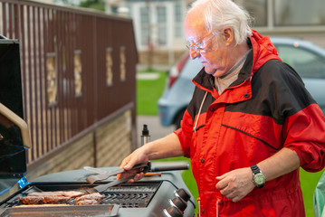 Elderly man in a red coat cooking meat on a BBQ