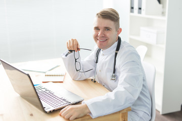 Healthcare, medical and people concept - Smiling doctor sitting at table with laptop and looking at the camera
