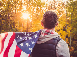 Attractive, stylish man holding a waving US Flag on the background of the rays of the setting sun and yellow trees. View from the back. Preparing for the holidays