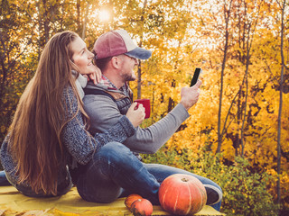 Attractive man and stylish woman holding a mobile phone and sitting on the terrace against the background of yellow trees and the setting sun. Happy relationship concept