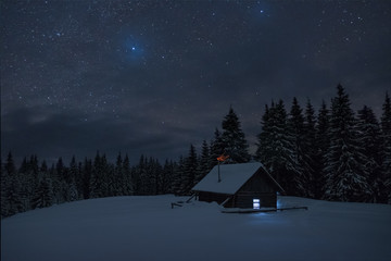Wooden cabin under stars. Lights shines through the window from inside of the house. Night landscape in winter.