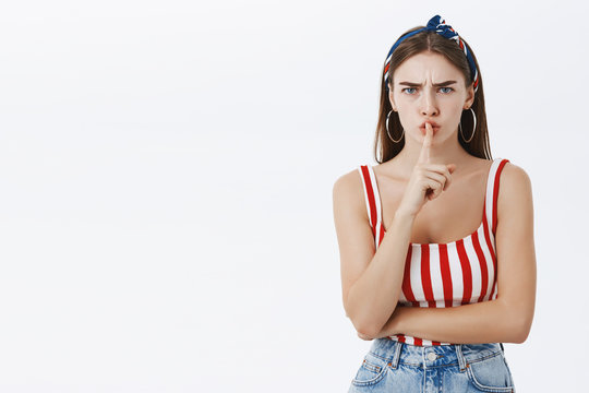 Shut up now. Irritated serious-looking bossy attractive woman in striped summer top and headband frowning while shushing at camera displeased and annoyed prohibiting speak over gray background