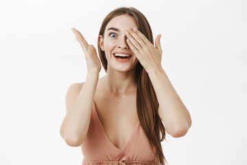 Excited woman cannot hold amazement inside covering eyes with palms peeking with one eye with surprise and joy being delighted seeing awesome gift or promotion against grey background