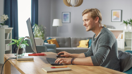 Portrait of the Handsome Man Working on Personal Computer while Sitting at His Desk. In the Background Stylish Cozy Living Room. Young Man Playing Computer Games.
