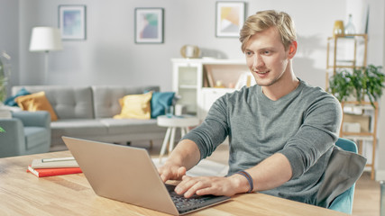 Fototapeta na wymiar Handsome Smiling Young Man Using Laptop While Sitting at the Desk of His Cozy Living Room.