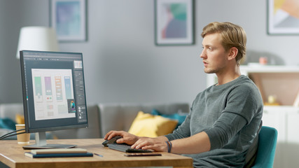 Handsome Focused Young Man Works on a UX / UI Mobile App Template, Uses Personal Computer while Sitting at His Desk in the Cozy Apartment.