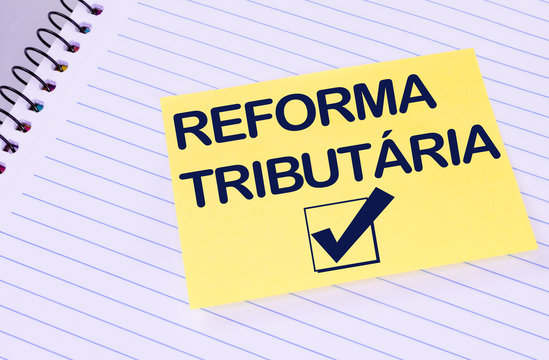 Sheet of yellow post note paper or self-adhesive notes written Tax Reform (in portuguese)with background notebook.