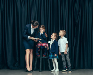 Cute smiling happy stylish children and female teacher on dark background. Beautiful stylish teen girls and boy standing together and posing on the school stage in front of the curtain. Classic style