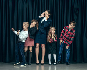Cute smiling happy stylish children and female teacher on dark background. Beautiful stylish teen girls and boy standing together and posing on the school stage in front of the curtain. Classic style