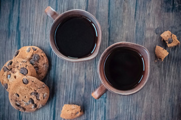 Chocolate cookies and two brown cups of black tea or coffee on brown on wooden background.