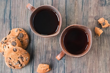 Chocolate cookies and two brown cups of black tea or coffee on brown on wooden background.