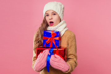blonde curly hairstyle teenager girl in knitted pink hat ,scarf and mittens mountain of gifts christmas box pink background studio. new Year present in hands of a female child making a wish copyspace