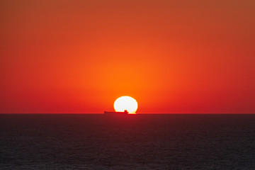Sunrise from sea water with passing ship on horizon. Red sun disc dawn above the ocean