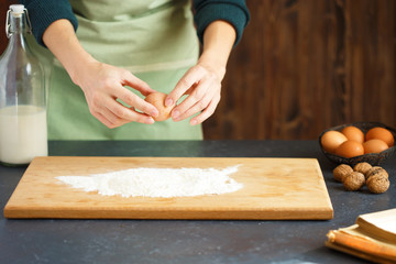 Women's hands knead the dough. The confectioner is driving an egg into the flour. On the wooden table are baking ingredients.