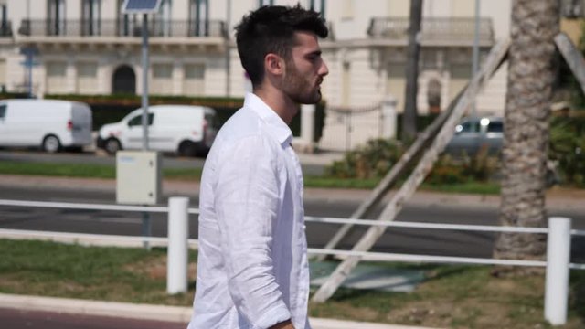 Attractive fit athletic young man soaking in the sun on seaside boardwalk or seafront, wearing white shirt open on naked torso, in Nice, France on the French Riviera