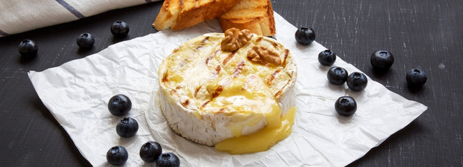 Grilled camembert cheese in paper with toasts, blueberries and walnuts on black background, side view. Close-up.