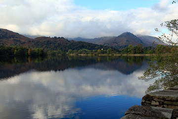 Fototapeta na wymiar Dow Bank, trees, and sky are reflected on to Grasmere water, Lake District, in the afternoon autumn sun. Foreground in sharp focus, with a slight softness in background to add character