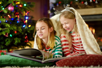 Happy little sisters reading a story book together by a fireplace in a cozy dark living room on Christmas eve.