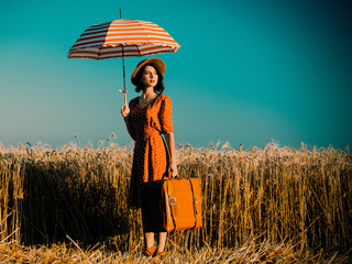 portrait of the beautiful young woman with red umbrella and brown suitcase standing on the field