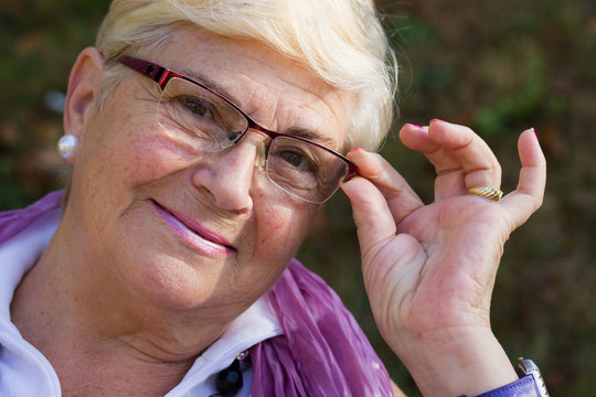 Closeup of smiling senior woman holding eyeglasses. Older blonde lady portrait looking at camera on sunny morning outdoors