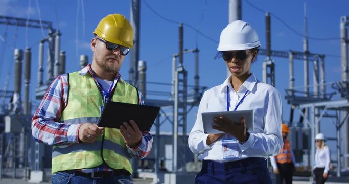 Medium shot of two electrical workers reviewing documents on a tablet during an inspection against the background of a transformer station