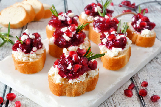 Holiday crostini appetizers with cranberries, pomegranates and feta cheese. Close up table scene on a white platter.