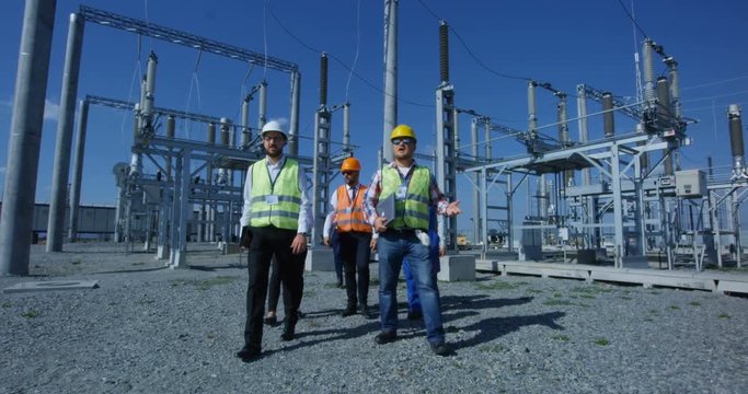 View of adult men in hardhat and vests walking with gadgets on transformer platform of solar electrical plant and talking