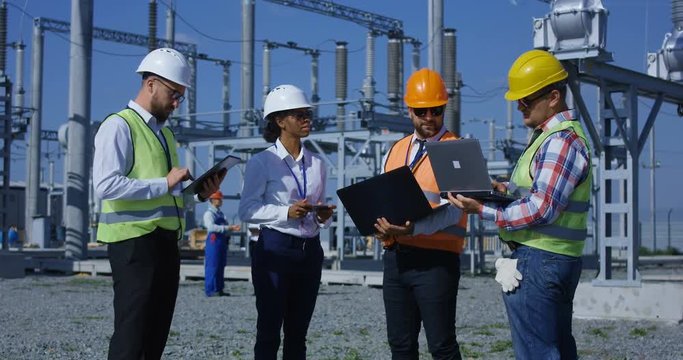 Medium shot of four electrical workers reviewing documents on a computer on a sunny day against the background of a transformer station