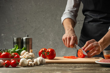 The concept of nutrition. The chef cuts tomatoes on the background of a concrete wall, with ingredients for cooking pizza, pizza sauce, sauce for Italian pasta - obrazy, fototapety, plakaty