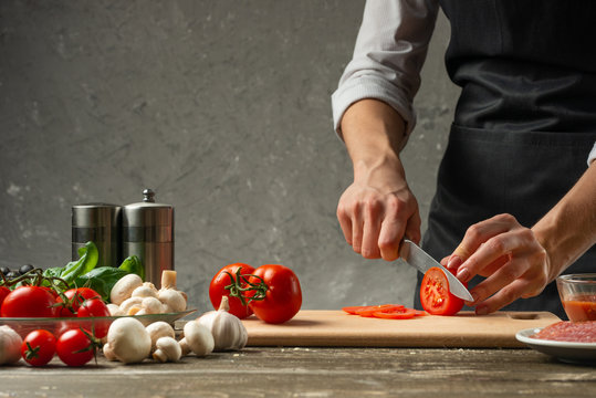 The concept of nutrition. The chef cuts tomatoes on the background of a concrete wall, with ingredients for cooking pizza, pizza sauce, sauce for Italian pasta