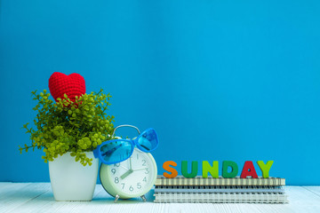 SUNDAY letters text and notebook paper, alarm clock and little decoration tree in white vase on wooden background, hello Sunday weekend concept.