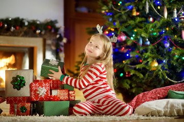 Happy little girl wearing Christmas pajamas playing by a fireplace in a cozy dark living room on Christmas eve.