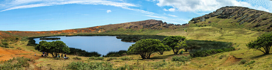 Vista panor&acirc;mica da cratera do vulc&atilde;o Rano Raraku, com um lago central, &aacute;rvores ao fundo e encostas montanhosas, capturada na Ilha de P&aacute;scoa em um dia ensolarado, destacando a natureza ex&oacute;tica