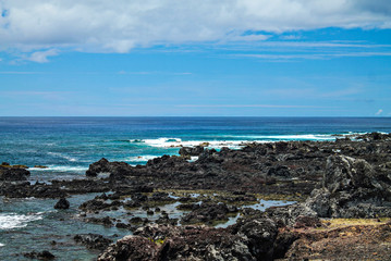 Costa rochosa da Ilha de Páscoa, com pedras em destaque no primeiro plano e o mar azul profundo ao fundo, em um dia ensolarado, capturando a beleza natural do local