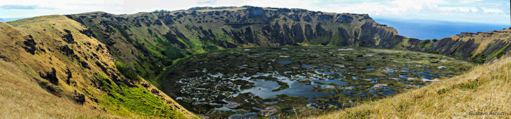 Vista panorâmica da cratera do vulcão Rano Kau, na Ilha de Páscoa, com encostas íngremes, vegetação ao redor e o céu azul ao fundo, destacando a beleza geológica e natural do local