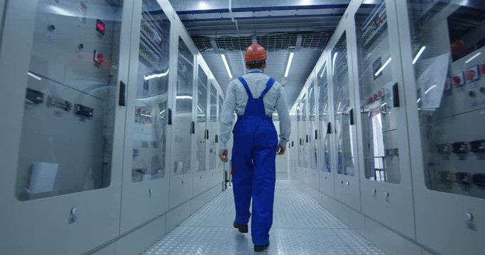 From below back view of Korean man in overall and hardhat walking in corridor with electrical equipment of solar power station