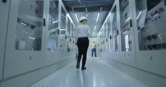 Back view of caucasian woman in hardhat walking in control center hallway of solar power station with colleagues