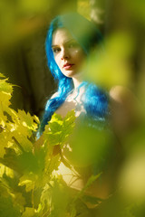 Schöne junge unkonventionelle Frau Emo mit individuellen blauen Haaren, schaut im Herbstwald zwischen grünen gelben Blättern hervor.