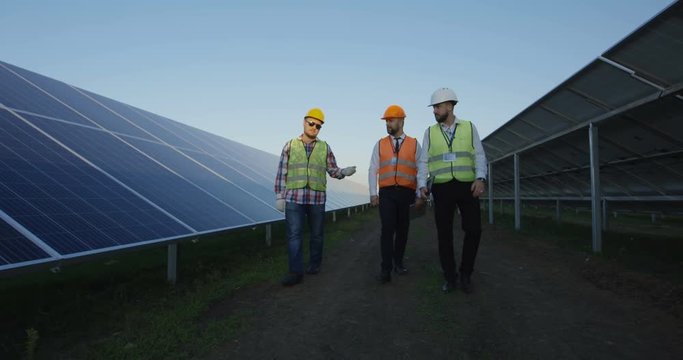 Group of adult men on field with rows of solar panels having professional discussion while working in team