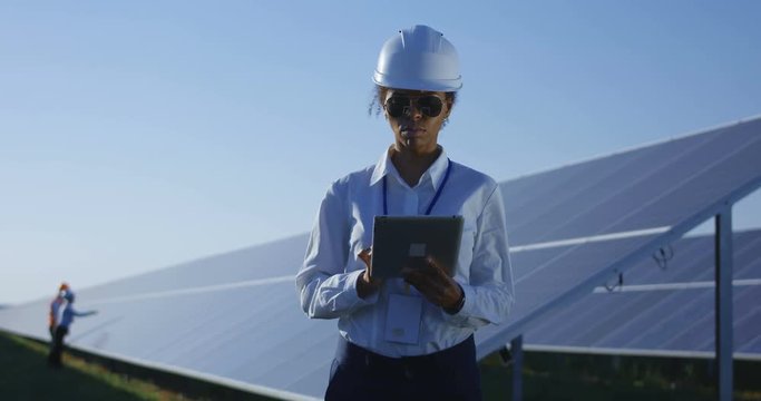Medium shot of a female electrical worker typing on her tablet inbetween long rows of photovoltaic solar panels