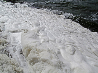 A lot of foam along the shore created by waves of water, close-up view. Summer scene of a lake or river. Concept of pollution of nature and a ecology problems of the environment.