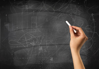 Female hand holding white chalk in front of a blackboard with scribbles and plans drawn on it 
