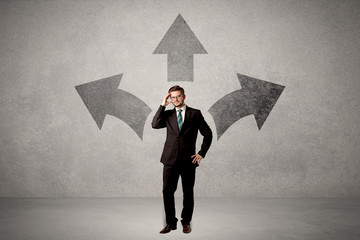 Charming businessman in doubt, choosing from three directions in front of a grey wall
