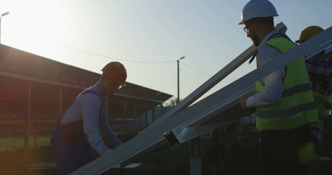 Medium shot of three workers in a uniform and hardhat installing photovoltaic panels on a metal basis on a solar farm